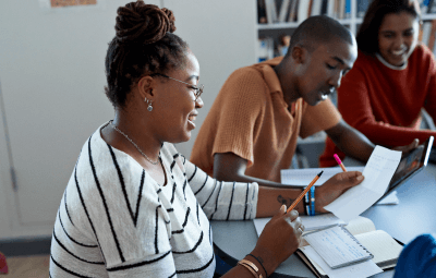 Side view of smiling young female student reading paper while sitting by friends at desk in library