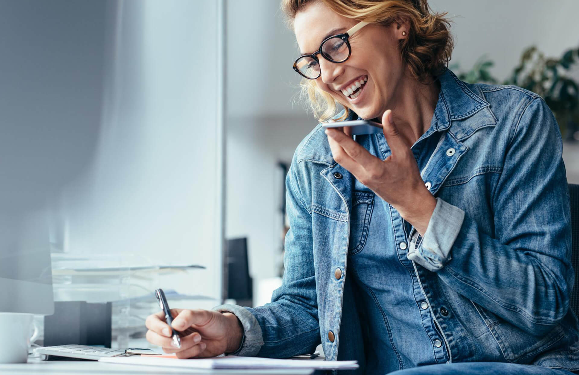 A woman laughing as she talks on the phone with someone while writing things down in a notebook