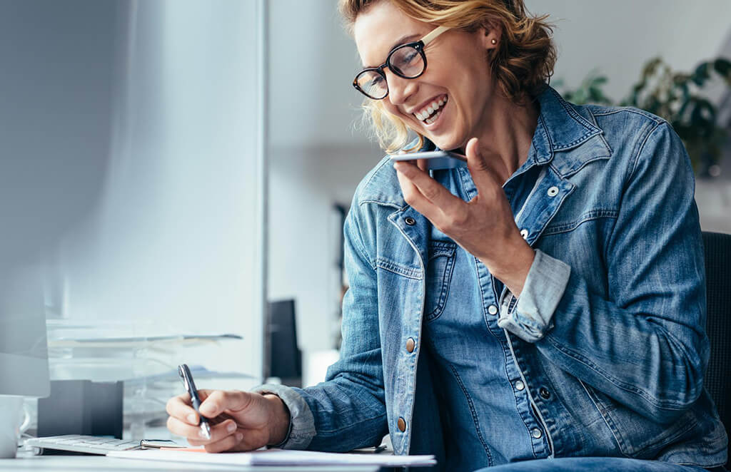 A woman laughing as she talks on the phone with someone while writing things down in a notebook