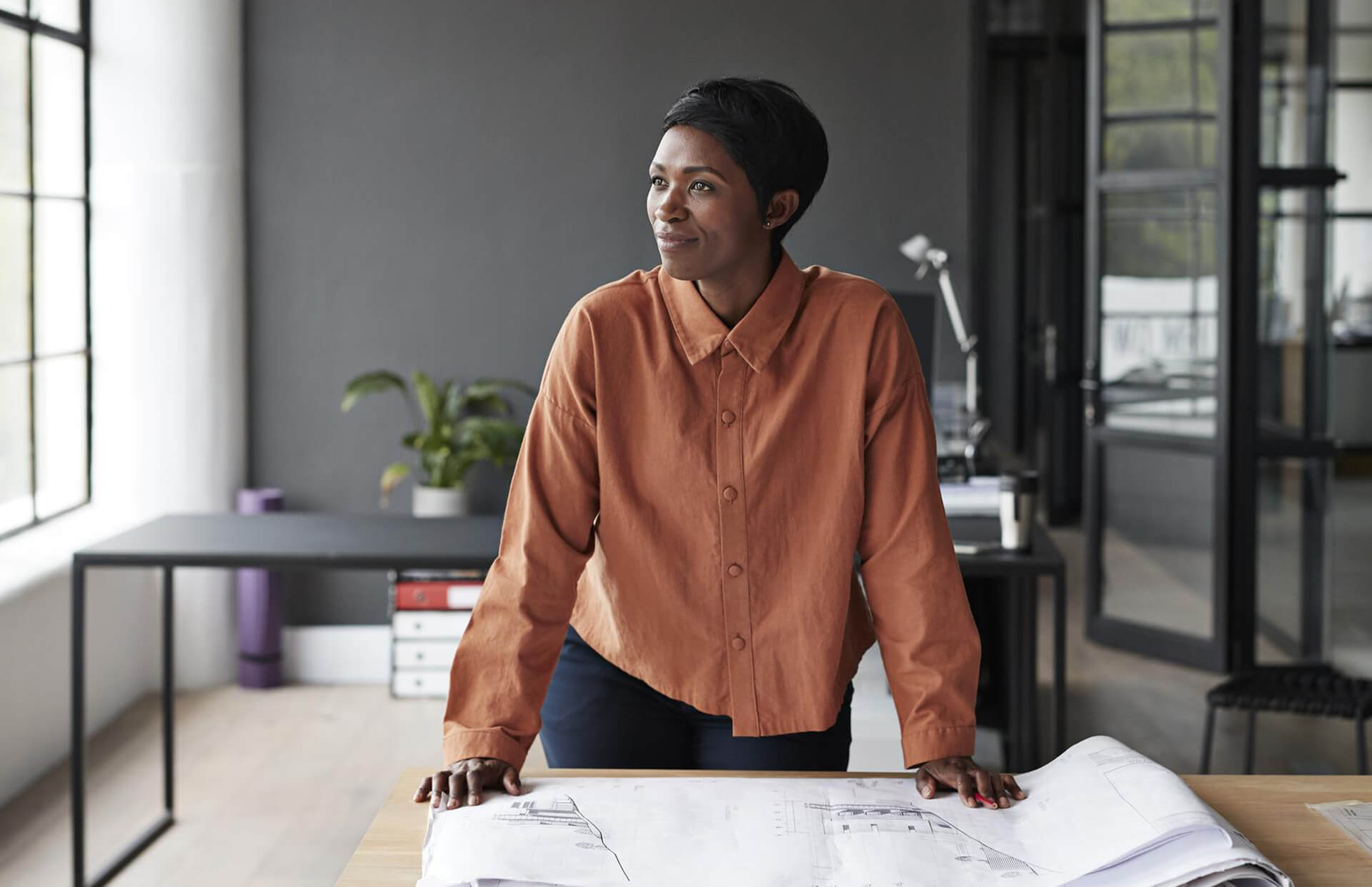 A woman standing at her desk looking over blueprints and gazing out of the window