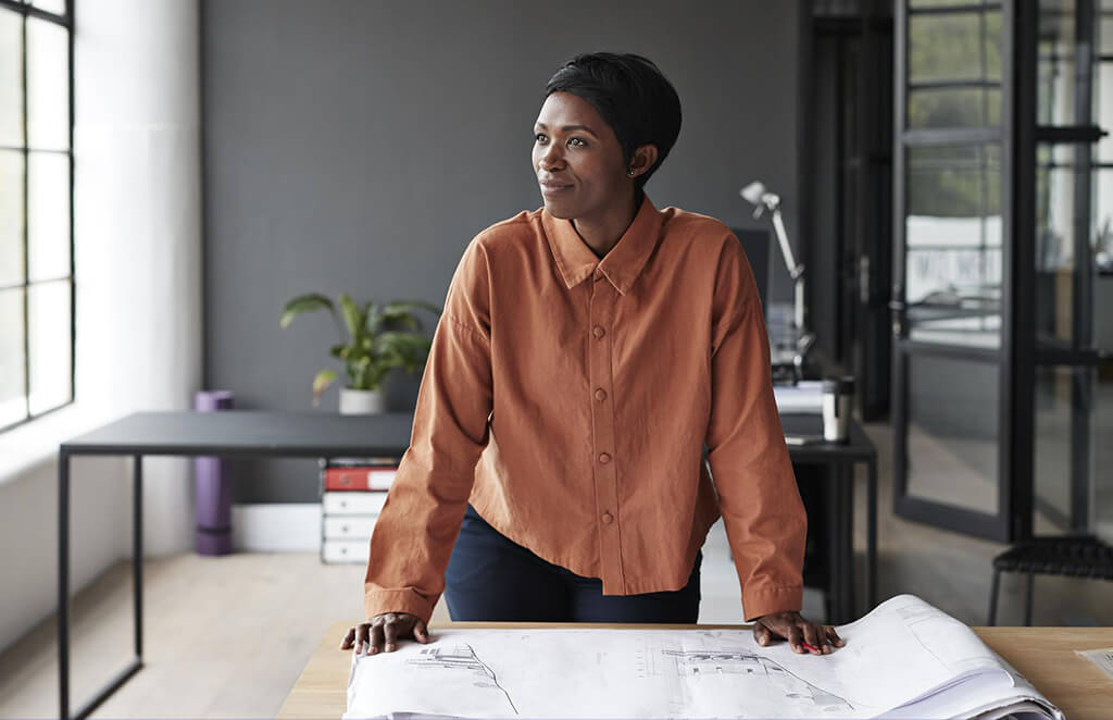A woman standing at her desk looking over blueprints and gazing out of the window