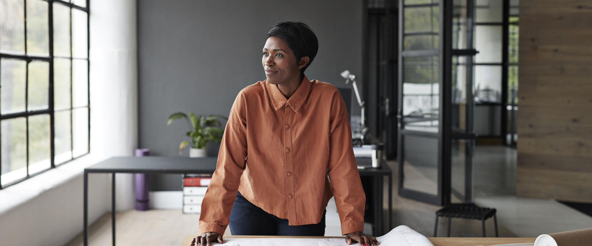 A woman standing at her desk looking over blueprints and gazing out of the window