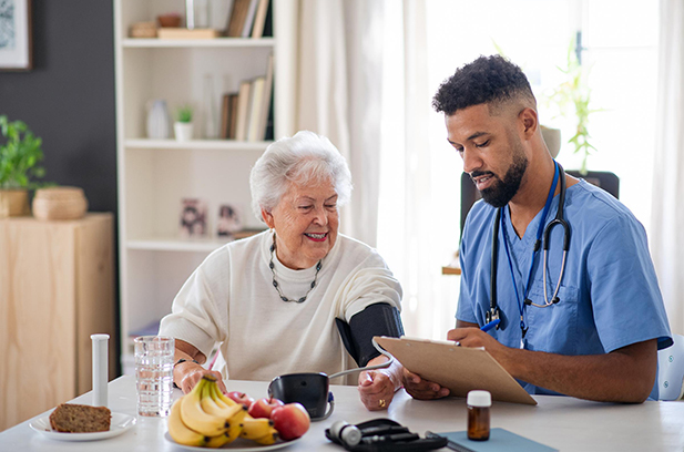 Elderly woman smiling as a healthcare professional in scrubs checks her blood pressure and reviews notes at a home setting. Items like fruit, bread, and medication are on the table.